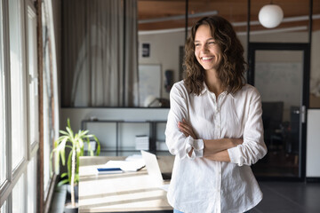 Cheerful young business professional woman in white casual shirt standing in office with arms...