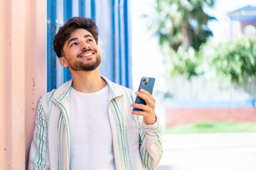 Handsome Arab man using mobile phone at outdoors looking up while smiling