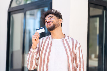 Handsome Arab man at outdoors holding a credit card and thinking