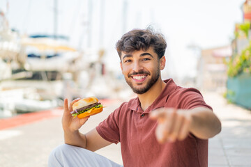 Young Arabian handsome man holding a burger at outdoors points finger at you with a confident...
