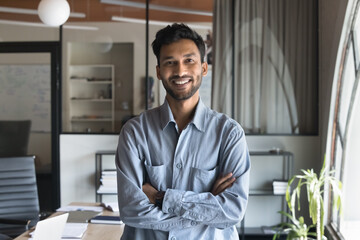 Happy successful attractive Indian business man in casual standing in office with arms crossed, looking at camera with toothy smile, posing for professional portrait with workplace in background - obrazy, fototapety, plakaty