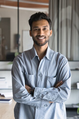 Cheerful handsome Indian businessman standing in office with hands folded, looking at camera, smiling. Young entrepreneur man, business professional, manager vertical portrait - obrazy, fototapety, plakaty