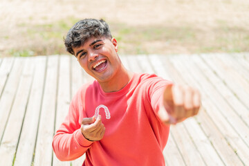 Young hispanic man holding invisible braces at outdoors points finger at you with a confident...