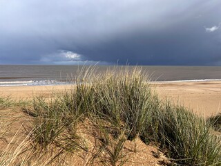 Man looking to ocean horizon beautiful sandy beach landscape grey stromy sky clouds and from grassy banks of sand dunes rolling waves on flat sea on Spring day in Winterton Norfolk East Anglia uk 