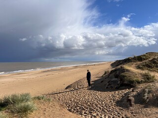 Man looking to ocean horizon beautiful sandy beach landscape grey stormy sky clouds and from grassy banks of sand dunes rolling waves on flat sea on Spring day in Winterton Norfolk East Anglia uk 