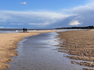 Stunning beautiful coast landscape of white sandy beach at low tide inlet river on shore to horizon with couple walking blue sky calm sea and white cloud Spring cold fresh day at Winterton Norfolk uk 
