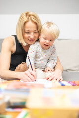 Obraz na płótnie Canvas Caring young Caucasian mother and small son drawing painting in notebook at home together. Loving mom or nanny having fun learning and playing with her little 1,5 year old infant baby boy child