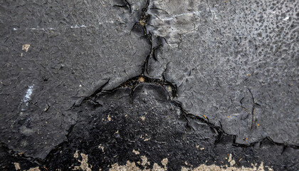 A close-up black and white shot of a cracked wall showing signs of deterioration and wear. The cracks cut through the concrete surface, creating a striking contrast between the dark shadows and light