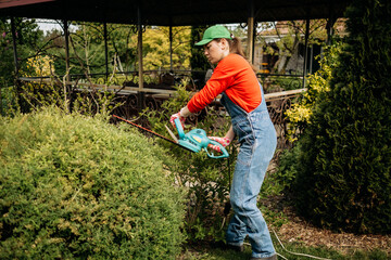A woman gardener in work uniform trims a bush with electric scissors. Handmade in the summer season.