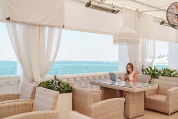 Woman coffee cafe laptop sea. Female freelancer using laptop on coffee break in modern cafe with sea view in the morning