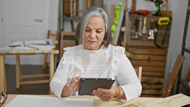 Elderly woman using tablet in a woodwork workshop surrounded by tools and workbenches