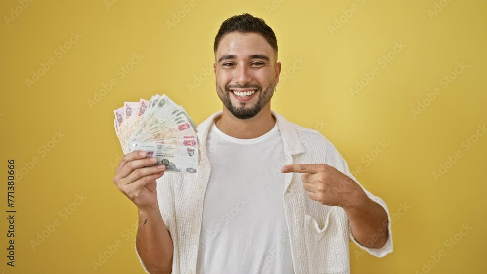 Canvas Prints Young, confident arab man smiling joyfully, holding uae banknotes, pointing, with hand and finger, isolated on yellow background