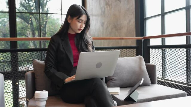 The young Asian businesswoman is working on her laptop, referencing business data from her tablet at the coffee shop