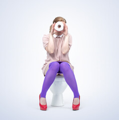 A young woman is sitting on the toilet and looking through a roll of toilet paper, isolated on a light blue background