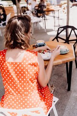 Charming woman in a restaurant, cafe on the street. She sits at the table and eats a cake with a fork. Dressed in a red sundress with white polka dots.