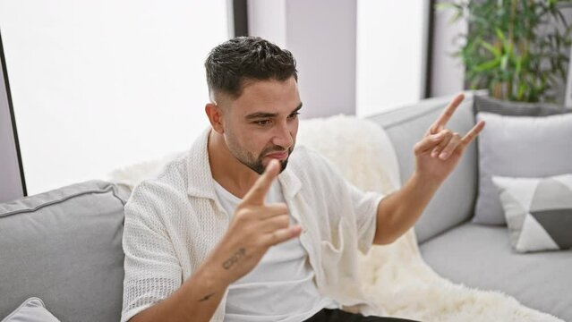 Hipster arab man rocking out on his living room sofa, hands raised in iconic rock symbol, letting loose his inner music star in a burst of wild, heavy metal expression