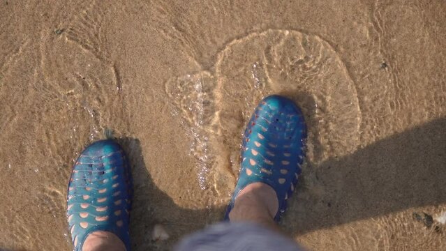 Wave washes feet of man in aquashoes on sandy beach. Modern protective silicone shoes on legs in sea on sunny day close up. POV top view of water flows from ocean waves.