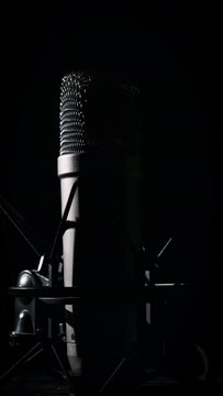 Vertical, studio microphone rotates on a black background in backlight close-up. Condenser microphone with chrome grid on surface. Concept recording studio, voice, podcast, karaoke. Copy space