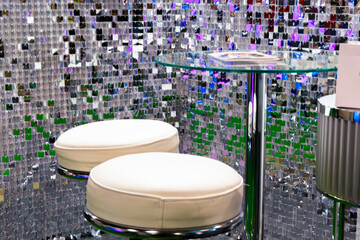 Two white bar stools and a glass tabletop on a silver shiny background of small foil squares. There is a disco ball hanging above the bar