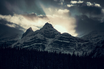 Snow-covered peaks with clouds hovering above and trees in the foreground.