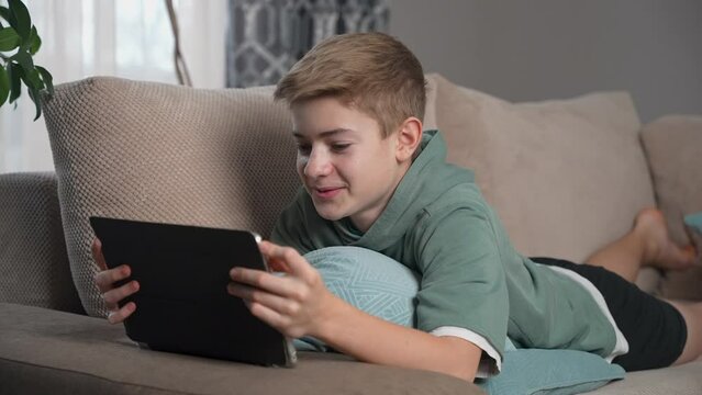Teenage Boy lying on sofa using tablet. Scrolling social media, surfing internet enjoying time at home alone. Child sitting on coach in living room with device. Listen to music, watching videos, smile