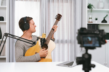 Fototapeta na wymiar Host channel in smart singer recording by camera, playing guitar along singing, broadcasting on social media channel, wearing headphones to record video streamer at white modern studio. Pecuniary.