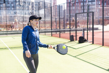 A girl in sportswear is training on a paddle tennis court. The girl is hitting the ball against the glass to make a rebound. Concept of women playing paddle.