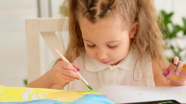 a happy and joyful child paints at a table in a bright room, a little girl learns to draw with a brush and gouache, a drawing lesson, close-up