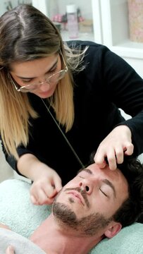 Vertical picture of focus beautician doing male eyebrow hair removal by threading technique. She is a epilation specialist in a beauty center. Concept wellness and body care