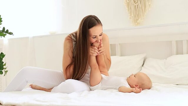 mom and baby are playing with their legs kissing them and having fun together in the children's bright room on the bed, happy motherhood with maternity leave, mom and baby, maternal love