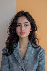 Obraz premium A portrait of a young woman with long, wavy hair and subtle makeup, wearing a smart gray blazer. She stands against a dual-tone background