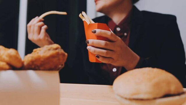 Closeup image of a woman holding and eating french fries and hamburger with fried chicken on the table at home