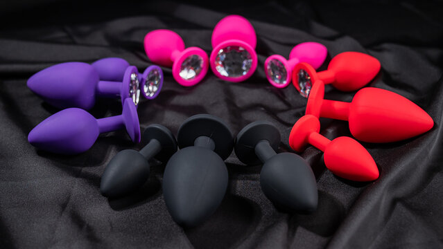 A set of silicone anal plugs in different colors and sizes on a black silk sheet. 
