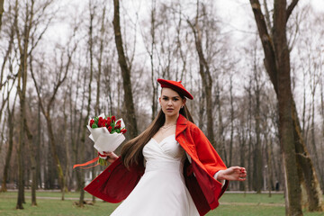 Young woman 20s in red french beret and jacket dressed white dress, walking in city park with...