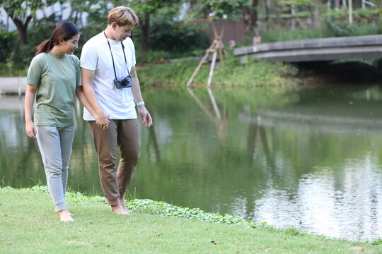 man couple holds a camera to take photos of his Asian female friend and takes pictures of the scenery while walking and relaxing in the garden. The couple walks barefoot on the grass in the garden.