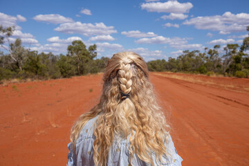 Blonde woman with braids looking down a red dirt country road into the distance in the arid...