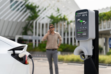 Young man recharge EV electric vehicle at green city commercial mall parking lot while talking on...