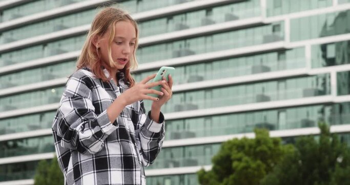 Teenage girl socializing on smart phone smart phone. Teenager is standing against modern building in city. She is wearing checkered pattern shirt.