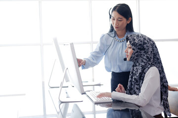Smiling Asian and Muslim women are colleagues and call center or secretary operator with headset and a microphone together. They are Consulting to solve problems about customers at office
