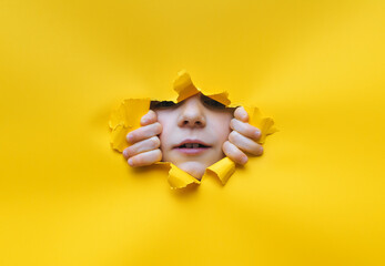 A little girl looks into a torn hole in yellow paper. Hands, nose and mouth. The concept of...