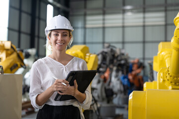 Female engineer in a hard hat using a digital tablet with industrial robotic arms in the background inside a manufacturing plant.
