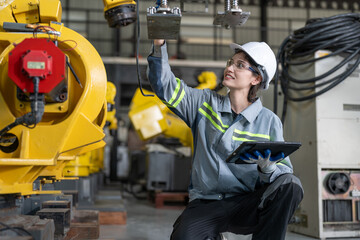 Female industrial engineer in hard hat inspecting new robot arms machinery components while holding a digital tablet in a manufacturing plant.