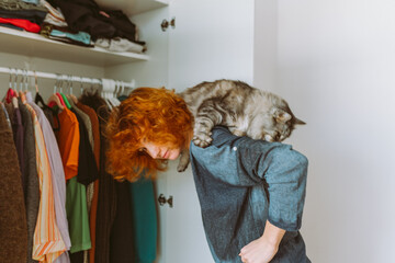 red-haired teenage girl and big cat choose things from closet