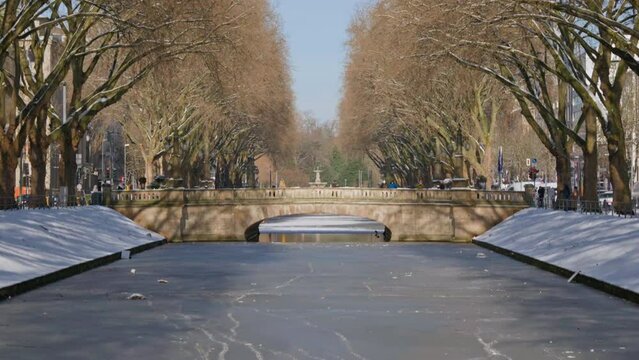 Clear winter view of bridge over frozen canal flanked by leafless trees