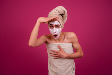 A scared man with a towel on his head and a mask of cream on his face. Pink background.