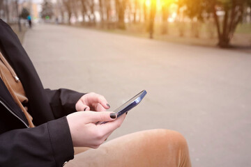 A girl sits on a bench in a city park with a smartphone in her hands.