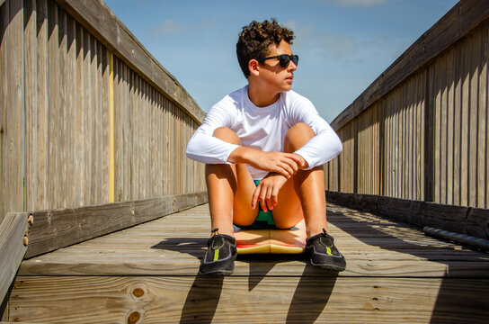Teenage male surfer wearing sunglasses and sitting on his surfboard relaxing 