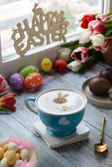 A cup of latte coffee against window toped with mousse of milk ,latte art draw in bunny shape and festive Easter decoration with chocolate Easter eggs and tulip flowers with spoon