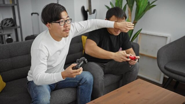 Two asian men intensely playing with video game controllers in a cozy living room, displaying leisure and technology.