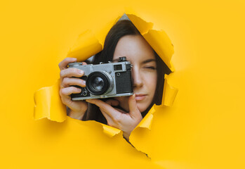 A young paparazzi girl with a rare SLR camera looks out from her hiding place and carefully watches what is happening. Yellow paper hole. Tabloid press. Looking for a subject for stock photos. - obrazy, fototapety, plakaty